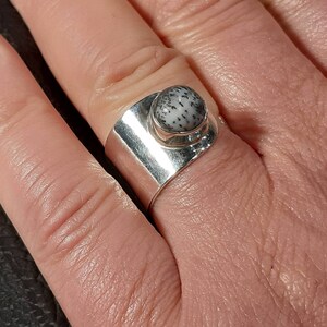 May include: A silver ring with a white and black stone set in a bezel setting. The ring is wide and has a flat band.