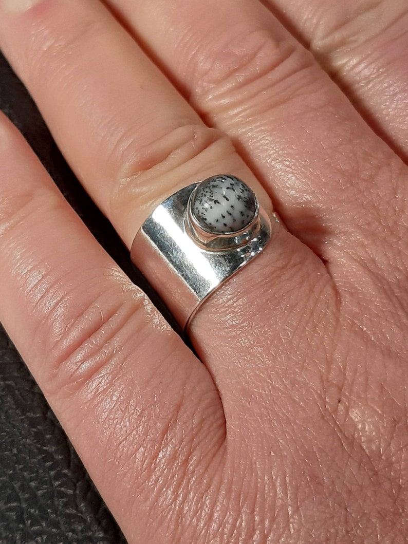 May include: A silver ring with a white and black stone set in a bezel setting. The ring is wide and has a flat band.