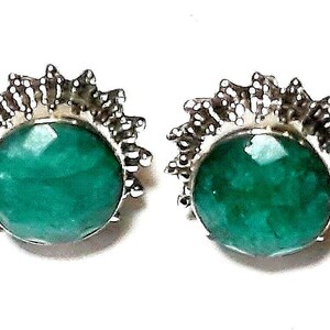 Puede incluir: Un par de pendientes de bot&oacute;n con grandes piedras preciosas redondas, facetadas, de color verde esmeralda. Las piedras preciosas est&aacute;n engastadas en un metal plateado con un dise&ntilde;o de rayos de sol puntiagudos alrededor de los bordes. Sobre fondo blanco.