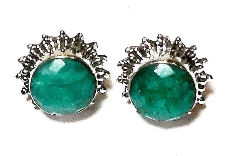 Puede incluir: Un par de pendientes de bot&oacute;n con grandes piedras preciosas redondas, facetadas, de color verde esmeralda. Las piedras preciosas est&aacute;n engastadas en un metal plateado con un dise&ntilde;o de rayos de sol puntiagudos alrededor de los bordes. Sobre fondo blanco.