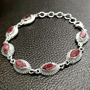 May include: A silver bracelet with a chain link design and six oval-shaped red gemstones. The gemstones are set in silver bezels and have a textured surface.