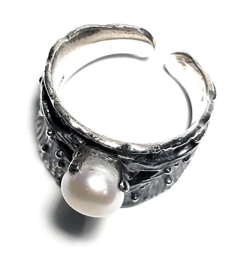 May include: A silver ring with a textured band and a white pearl. The ring is open and adjustable.