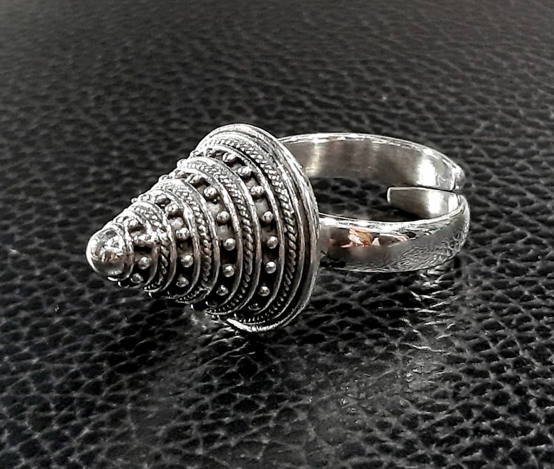 May include: A silver ring with a cone-shaped design. The cone is decorated with a series of concentric circles and small dots.