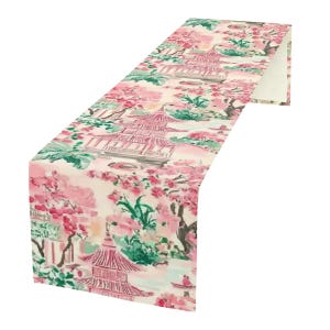 May include: A pink and green table runner with a Japanese garden design. The runner features a pagoda, cherry blossoms, and other floral elements.