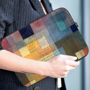 May include: A rectangular laptop sleeve featuring a colorful, abstract geometric design with a black zipper. The artwork incorporates a variety of colors, including blues, greens, browns, and yellows, creating a unique and artistic appearance. The sleeve is held by a person.