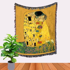 May include: A woven tapestry throw blanket featuring a reproduction of Gustav Klimt's "The Kiss" painting. The artwork depicts a couple embracing in a golden, mosaic-like style.