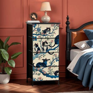May include: A four-drawer chest with a black frame and a cream-colored front panel. The panel features a repeating pattern of black and blue cats perched on flowering branches. The chest is topped with a black surface and sits on black legs.