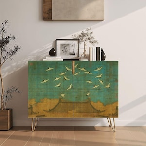 Puede incluir: Un mueble rectangular de dos puertas con una escena pintada en azul verdoso y dorado de grullas blancas volando sobre una estructura de techo tradicional. El mueble se apoya sobre cuatro patas de metal fino en forma de horquilla doradas.