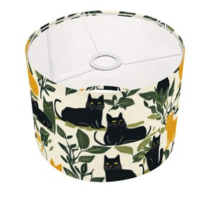 May include: A cylindrical lampshade featuring a cream-colored background with a repeating pattern of black cats and dark green foliage. The design includes scattered yellow shapes and a white interior frame with a central metal ring for mounting.