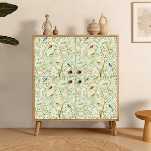 May include: A wooden cabinet with four doors featuring a light green background patterned with brown tree branches, green leaves, small orange flowers, and various colorful birds. The cabinet stands on four tapered wooden legs and has small round knobs.