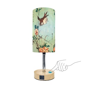 May include: A table lamp with a light green cylindrical shade featuring a Japanese-inspired floral and bird design. The lamp has a wooden base with a touch switch and a built-in outlet.