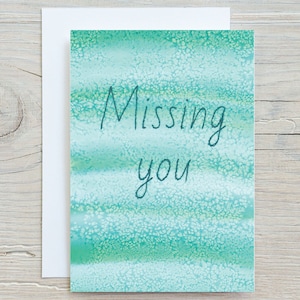 May include: A teal blue card with a white envelope. The card has a watercolour background and the text "Missing you" written in black.