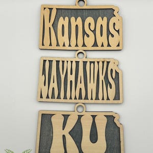 May include: Three wooden ornaments with a Kansas theme. The top ornament reads "Kansas", the middle one reads "JAYHAWKS", and the bottom one reads "KU". Each ornament has a light wood color with a dark gray background and a small hole for hanging.
