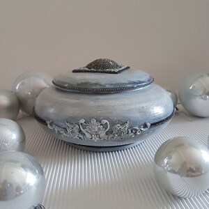 Op de afbeelding: Een decoratieve, lichtgrijze sieradendoos met deksel, versierd met sierlijke details en een kralen bovenkant. Zilveren kerstballen omringen de doos, wat een feestelijke sfeer cre&euml;ert. De doos heeft een gedetailleerd, decoratief ontwerp.