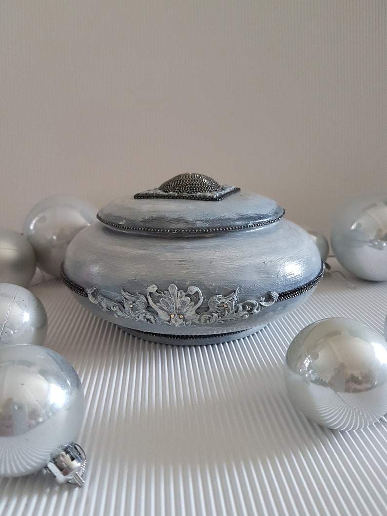 Op de afbeelding: Een decoratieve, lichtgrijze sieradendoos met deksel, versierd met sierlijke details en een kralen bovenkant. Zilveren kerstballen omringen de doos, wat een feestelijke sfeer cre&euml;ert. De doos heeft een gedetailleerd, decoratief ontwerp.