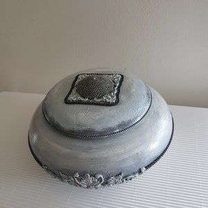 Op de afbeelding: Een decoratieve, zilver geverfde, ronde doos met een deksel. Het deksel heeft een vierkante, met kralen bezette versiering. De doos heeft een kralenrand en sierlijke, zilverkleurige bloemmotieven aan de basis. De doos staat op een wit oppervlak.