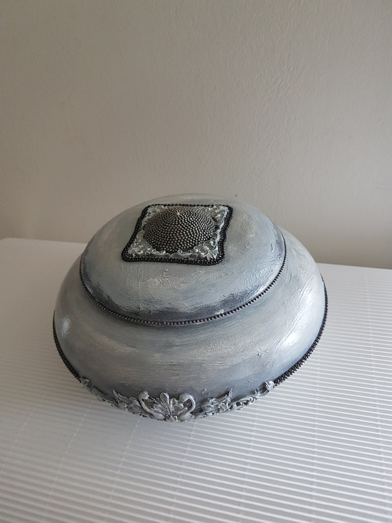 Op de afbeelding: Een decoratieve, zilver geverfde, ronde doos met een deksel. Het deksel heeft een vierkante, met kralen bezette versiering. De doos heeft een kralenrand en sierlijke, zilverkleurige bloemmotieven aan de basis. De doos staat op een wit oppervlak.