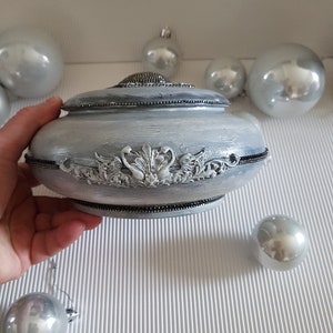 Op de afbeelding: Een zilverkleurige decoratieve doos met een deksel, met sierlijke reli&euml;fdetails en een kralenrand. De doos wordt door een hand vastgehouden, met zilveren ballen op de achtergrond. De doos heeft een ronde vorm.