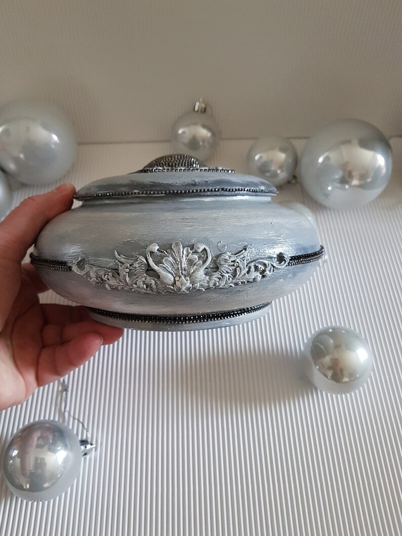 Op de afbeelding: Een zilverkleurige decoratieve doos met een deksel, met sierlijke reli&euml;fdetails en een kralenrand. De doos wordt door een hand vastgehouden, met zilveren ballen op de achtergrond. De doos heeft een ronde vorm.