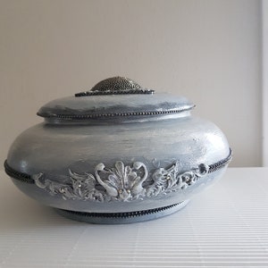 Op de afbeelding: Een decoratieve, lichtblauwe pot met deksel en zilverkleurige versieringen. De pot heeft een kralenrand en een getextureerde, metalen bovenkant. De voorkant van de pot heeft een gedetailleerd bloemenontwerp met kleine, sprankelende accenten.