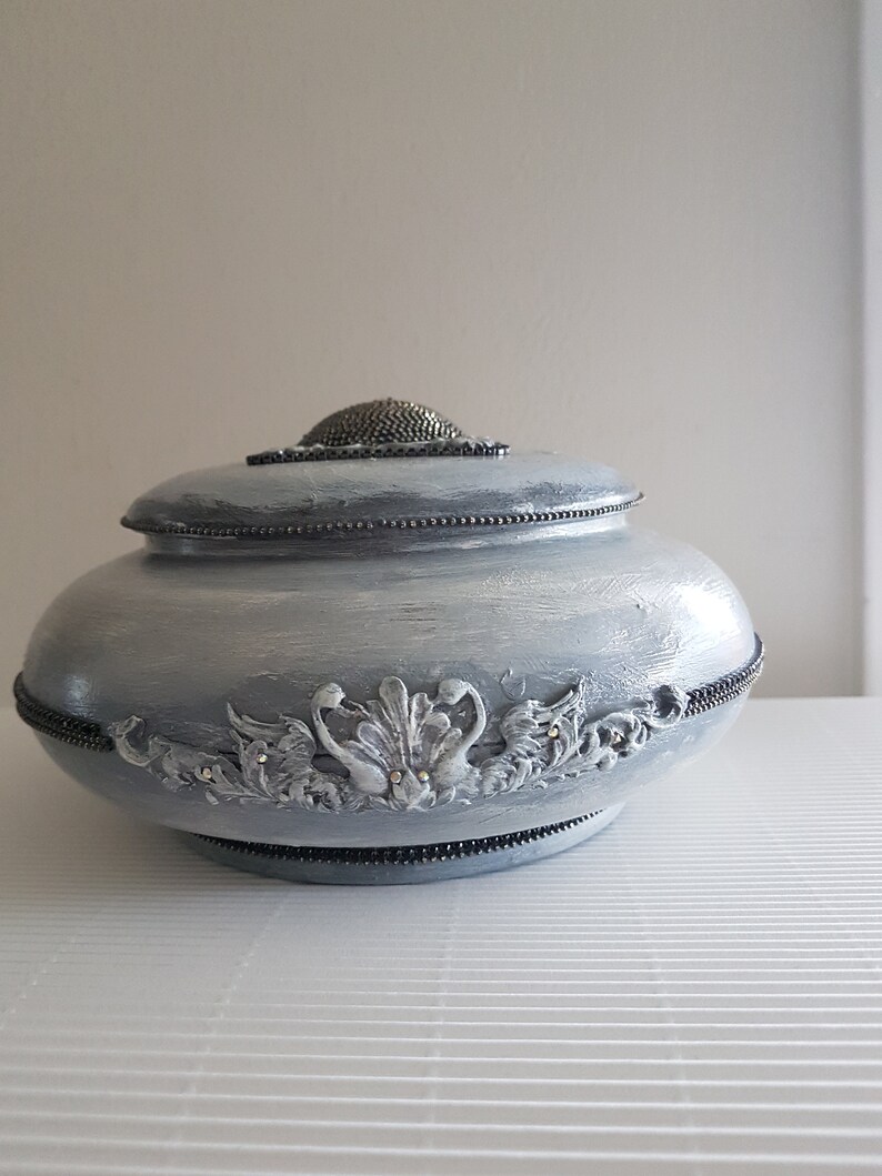 Op de afbeelding: Een decoratieve, lichtblauwe pot met deksel en zilverkleurige versieringen. De pot heeft een kralenrand en een getextureerde, metalen bovenkant. De voorkant van de pot heeft een gedetailleerd bloemenontwerp met kleine, sprankelende accenten.