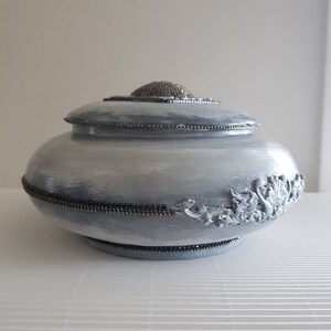 Op de afbeelding: Een lichtblauwe decoratieve urn met een deksel. De urn is versierd met zilverkleurige kralen en een decoratief bloemmotief aan de zijkant. Het deksel heeft een getextureerde zilverkleurige knop.