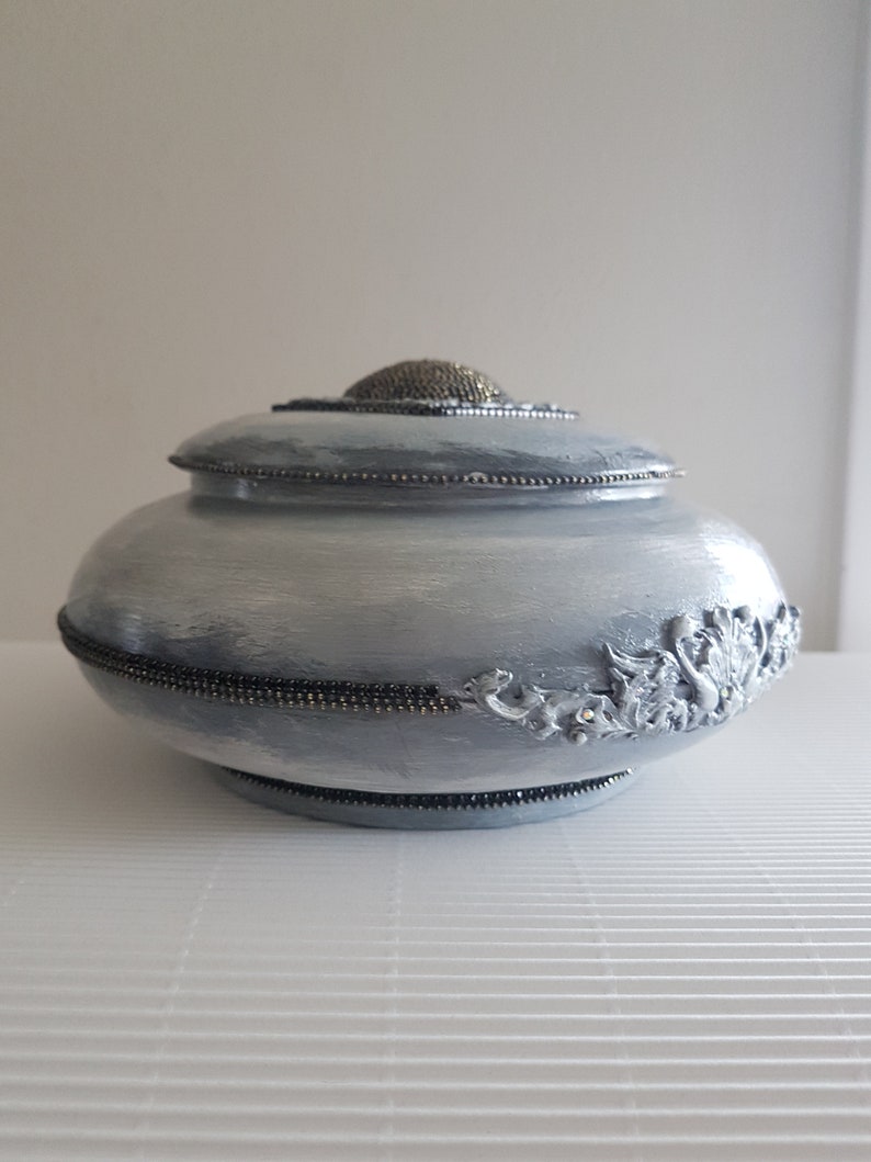 Op de afbeelding: Een lichtblauwe decoratieve urn met een deksel. De urn is versierd met zilverkleurige kralen en een decoratief bloemmotief aan de zijkant. Het deksel heeft een getextureerde zilverkleurige knop.