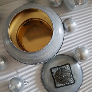 Op de afbeelding: Een zilver geschilderde, ronde container met een gouden interieur en een bijpassend deksel. Het deksel heeft een vierkant kralenontwerp. Verschillende zilveren kerstballen omringen de container en cre&euml;ren een feestelijke presentatie.