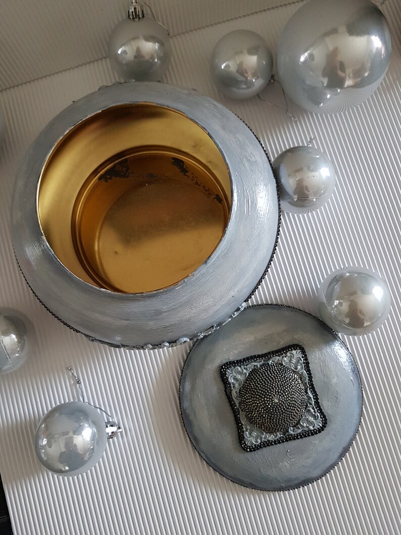 Op de afbeelding: Een zilver geschilderde, ronde container met een gouden interieur en een bijpassend deksel. Het deksel heeft een vierkant kralenontwerp. Verschillende zilveren kerstballen omringen de container en cre&euml;ren een feestelijke presentatie.