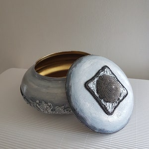 Op de afbeelding: Een ovale decoratieve doos met een deksel, geschilderd in grijstinten met een gouden binnenkant. Het deksel heeft een ruitvormig ontwerp van kralen. De basis van de doos heeft sierlijke, verhoogde bloemendetails.