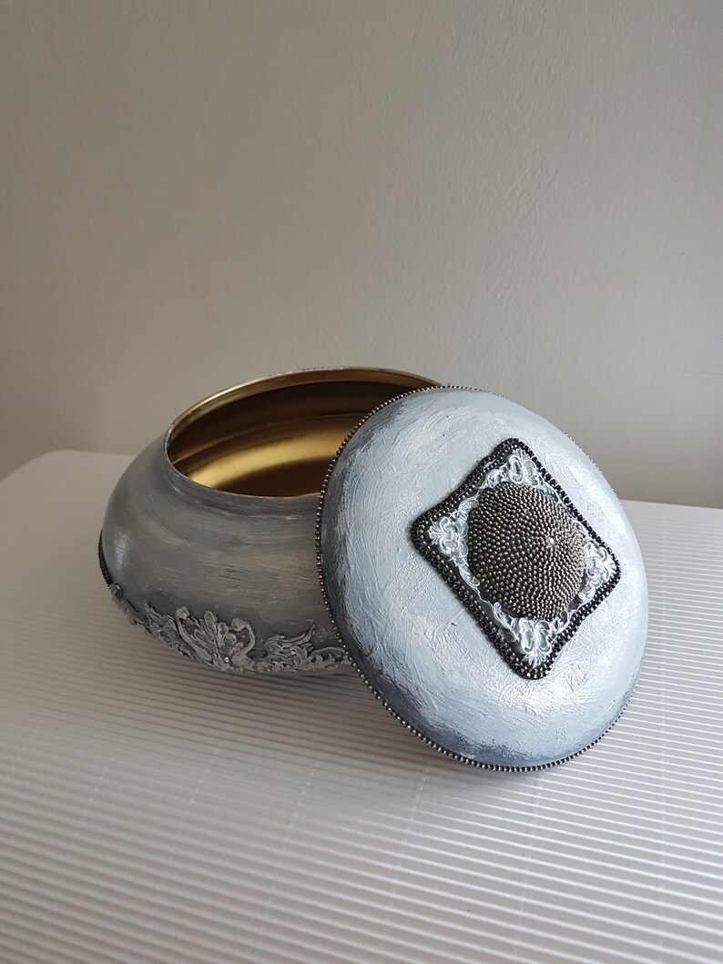 Op de afbeelding: Een ovale decoratieve doos met een deksel, geschilderd in grijstinten met een gouden binnenkant. Het deksel heeft een ruitvormig ontwerp van kralen. De basis van de doos heeft sierlijke, verhoogde bloemendetails.