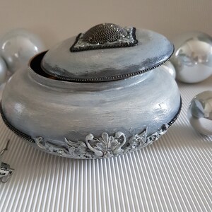 Op de afbeelding: Een decoratieve, lichtgrijze sieradendoos met een deksel. Het deksel heeft een kralenversiering en een zwarte rand. De doos heeft sierlijke zilverkleurige bloemendetails rond de basis en een kralenrand. Zilveren ornamenten omringen de doos.