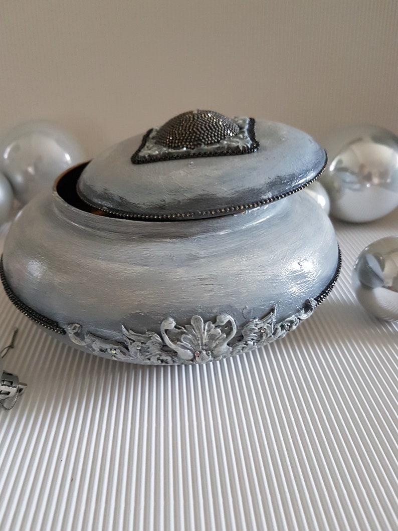 Op de afbeelding: Een decoratieve, lichtgrijze sieradendoos met een deksel. Het deksel heeft een kralenversiering en een zwarte rand. De doos heeft sierlijke zilverkleurige bloemendetails rond de basis en een kralenrand. Zilveren ornamenten omringen de doos.