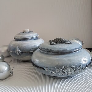 Op de afbeelding: Twee decoratieve, lichtblauwe potten met deksel en zilveren versieringen. De potten hebben een getextureerde, geverfde afwerking en zijn geaccentueerd met zilveren sierlijsten. Zilveren kerstballen staan op de achtergrond.