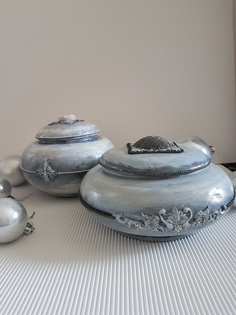 Op de afbeelding: Twee decoratieve, lichtblauwe potten met deksel en zilveren versieringen. De potten hebben een getextureerde, geverfde afwerking en zijn geaccentueerd met zilveren sierlijsten. Zilveren kerstballen staan op de achtergrond.