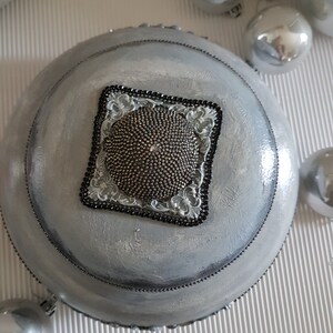 Op de afbeelding: Een zilver geschilderde, ronde decoratieve kom met een kralenrand en een vierkant, sierlijk middelpunt. Het middelpunt heeft een textuur, kralenontwerp omlijst door een zwarte kralenrand en decoratieve zilveren accenten. Zilveren kerstballen zijn verspreid.