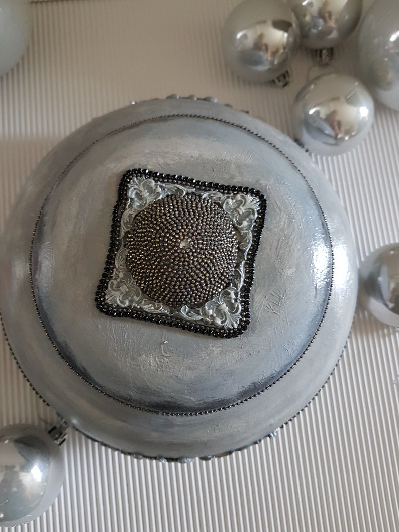 Op de afbeelding: Een zilver geschilderde, ronde decoratieve kom met een kralenrand en een vierkant, sierlijk middelpunt. Het middelpunt heeft een textuur, kralenontwerp omlijst door een zwarte kralenrand en decoratieve zilveren accenten. Zilveren kerstballen zijn verspreid.