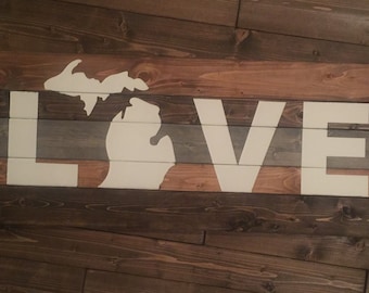 Könnte beinhalten: Ein Holzschild mit dem Wort "LOVE" in weiß auf einem gestreiften Hintergrund in Braun, Grau und Weiß. Der Bundesstaat Michigan ist ebenfalls in Weiß in der Mitte des Schildes gemalt.
