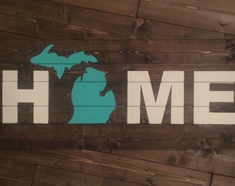 Könnte beinhalten: Ein Holzschild mit dem Wort "HOME" in weißen Buchstaben. Der Bundesstaat Michigan ist in Türkis in der Mitte des Schildes gemalt.