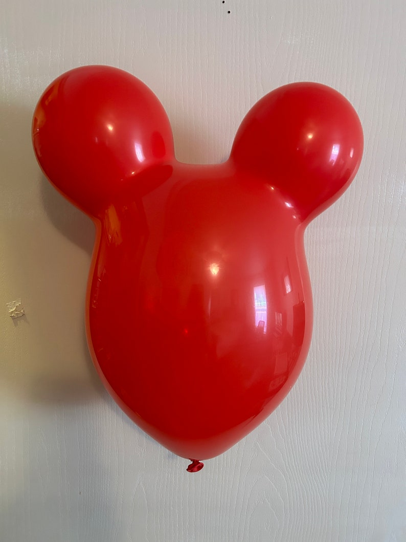 May include: A large, red balloon shaped like a cartoon mouse head. The balloon has two round ears on top and a rounded head shape. The balloon is inflated and appears to be made of a shiny, smooth material. The balloon is hanging against a white wall.