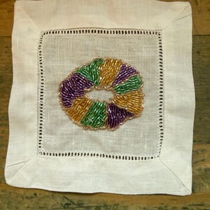 May include: A square white linen coaster with a beaded design. The beaded design is a circular shape with green, gold, and purple beads. The coaster has a stitched border.