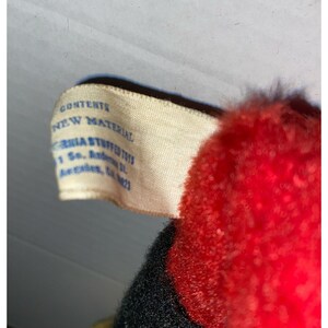 May include: A white tag with blue text that reads "Contents: New Material" and "California Stuffed Toys, 1 So. Antonio St., Arcadia, CA 91007". The tag is attached to a red and black stuffed toy.
