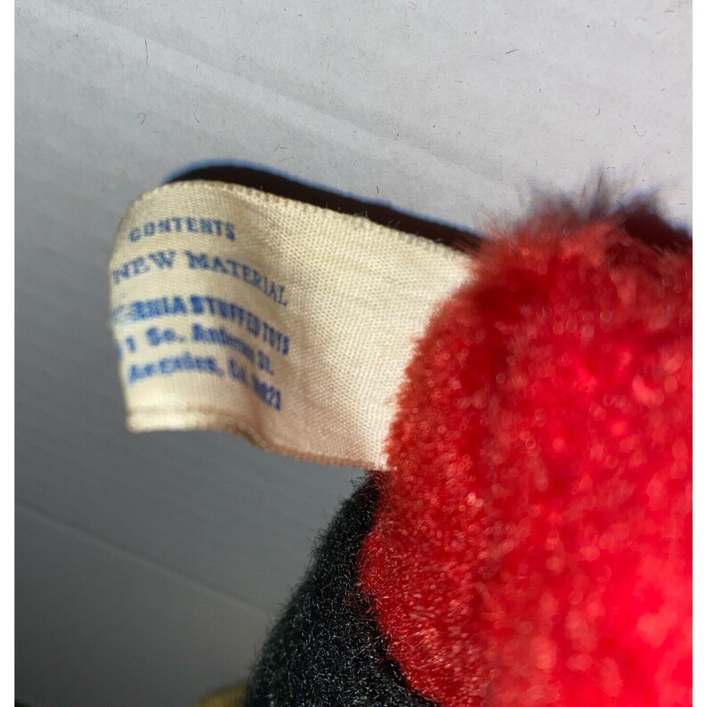 May include: A white tag with blue text that reads "Contents: New Material" and "California Stuffed Toys, 1 So. Antonio St., Arcadia, CA 91007". The tag is attached to a red and black stuffed toy.