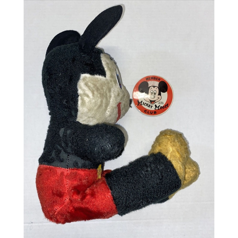 May include: Vintage Mickey Mouse plush toy wearing red shorts and a black and white outfit. The toy has a Mickey Mouse Club Member pin attached to its chest.