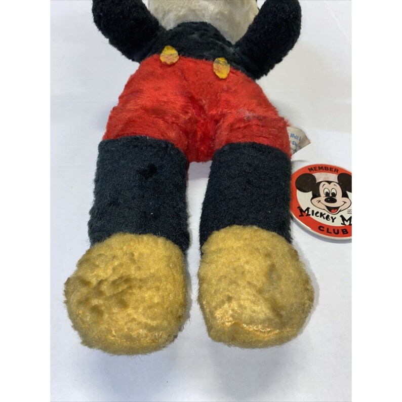 May include: Vintage plush Mickey Mouse toy wearing red shorts with yellow buttons, black shoes with yellow trim, and a black and white face. The toy has a Mickey Mouse Club Member tag attached to its leg.