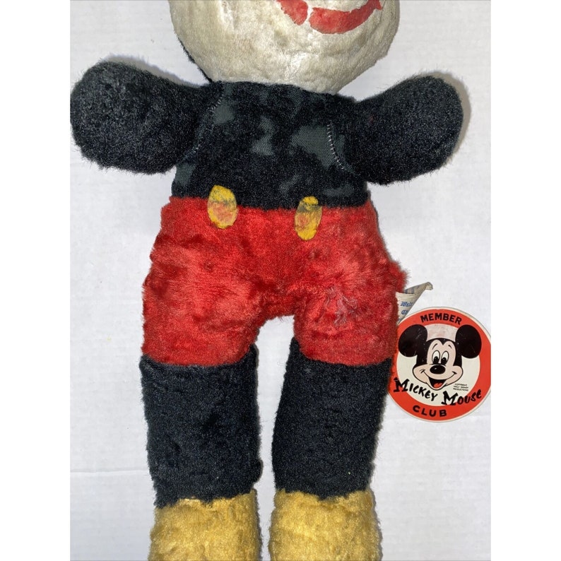 May include: Vintage plush Mickey Mouse toy wearing red shorts and black shoes. The toy has a Mickey Mouse Club member tag attached to its leg.