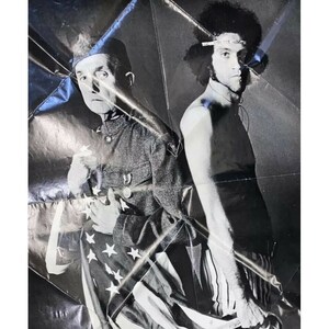 May include: Black and white photo of two men. One man is wearing a military uniform and holding an American flag. The other man is wearing a headband and a striped shirt.