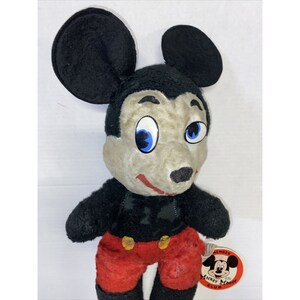 May include: Vintage plush Mickey Mouse toy with black fur, red shorts, and a black and white shirt. The toy has a grey face with blue eyes and a black nose. The toy has a tag that says "Member Mickey Mouse Club".