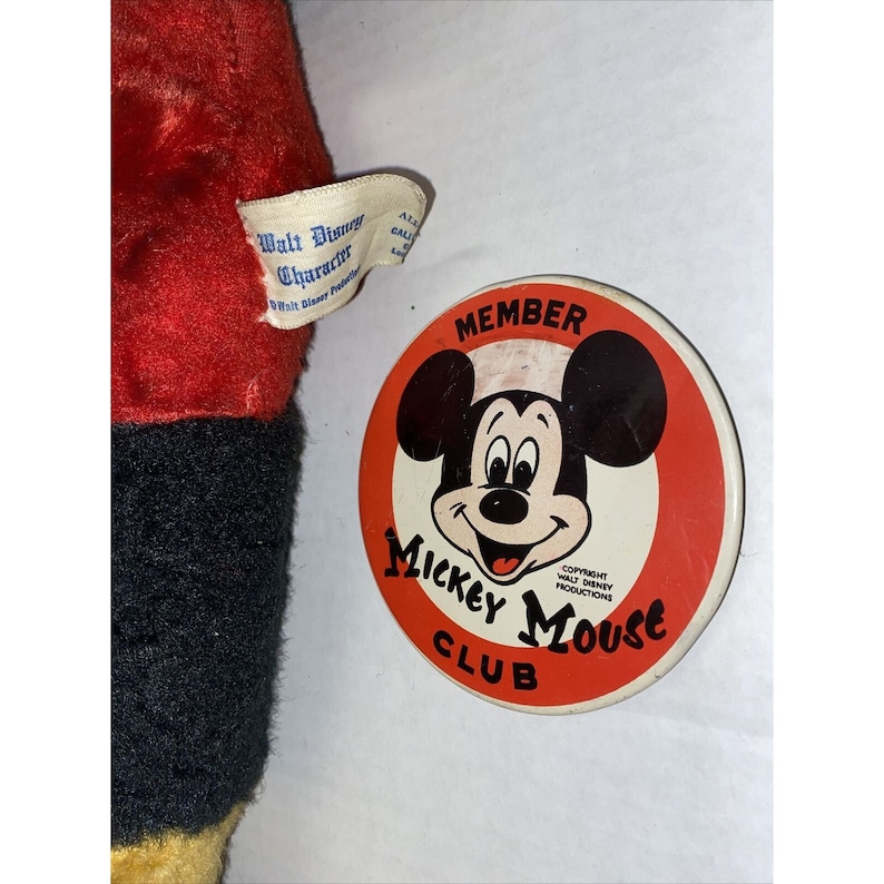 May include: A red, black and yellow plush Mickey Mouse toy with a Mickey Mouse Club member pin. The pin is red, white and black with a picture of Mickey Mouse's face and the text "MEMBER Mickey Mouse CLUB" and "COPYRIGHT WALT DISNEY PRODUCTIONS".