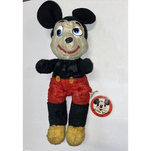 May include: Vintage Mickey Mouse plush toy with black fur, red trousers, and yellow shoes. The toy has a worn and faded appearance. It has a Mickey Mouse Club membership tag attached.